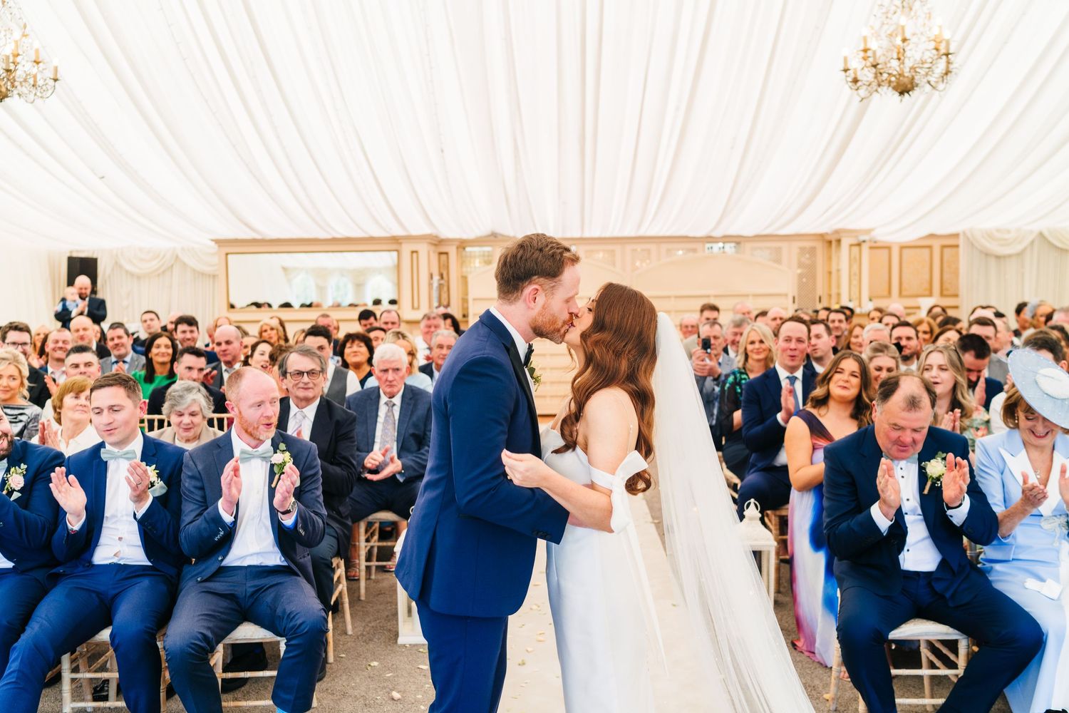 Castle Leslie WeddinNewlyweds share their first kiss at wedding reception while guests in blue suits applaud in elegant white tent setting.