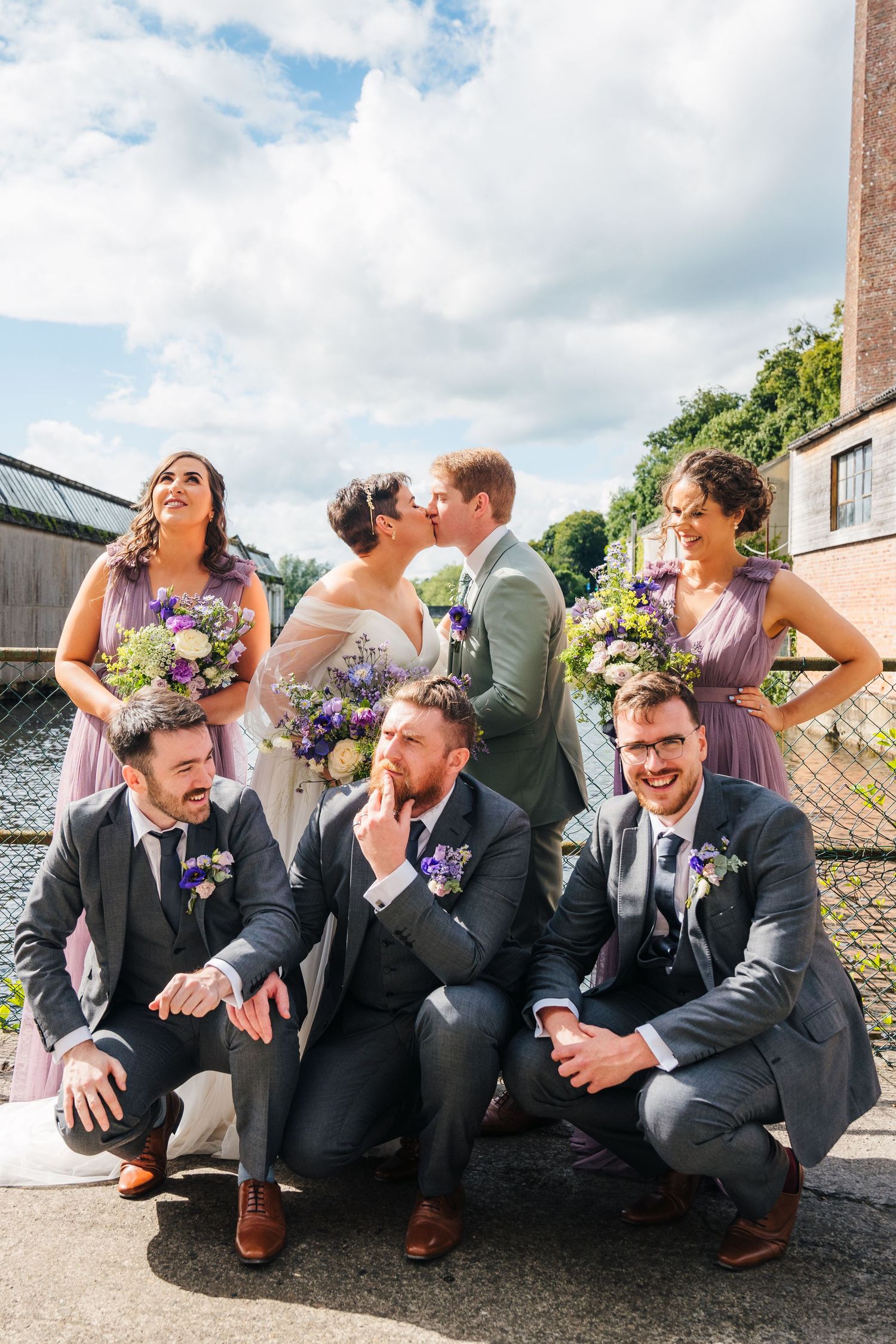 A wedding party in purple bridesmaids dresses and dark suits poses playfully outdoors on a sunny day.Millhouse slane wedding photographer du