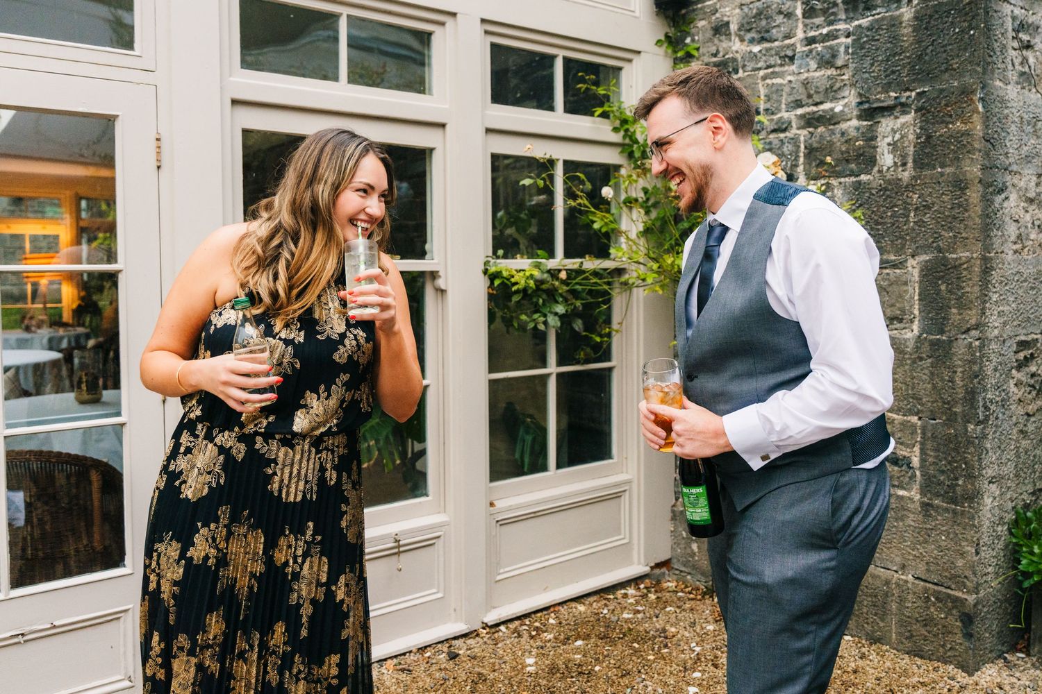 Millhouse slane wedding photographer duA pair of wedding guests laugh together while enjoying drinks outdoors near a stone wall and windows.