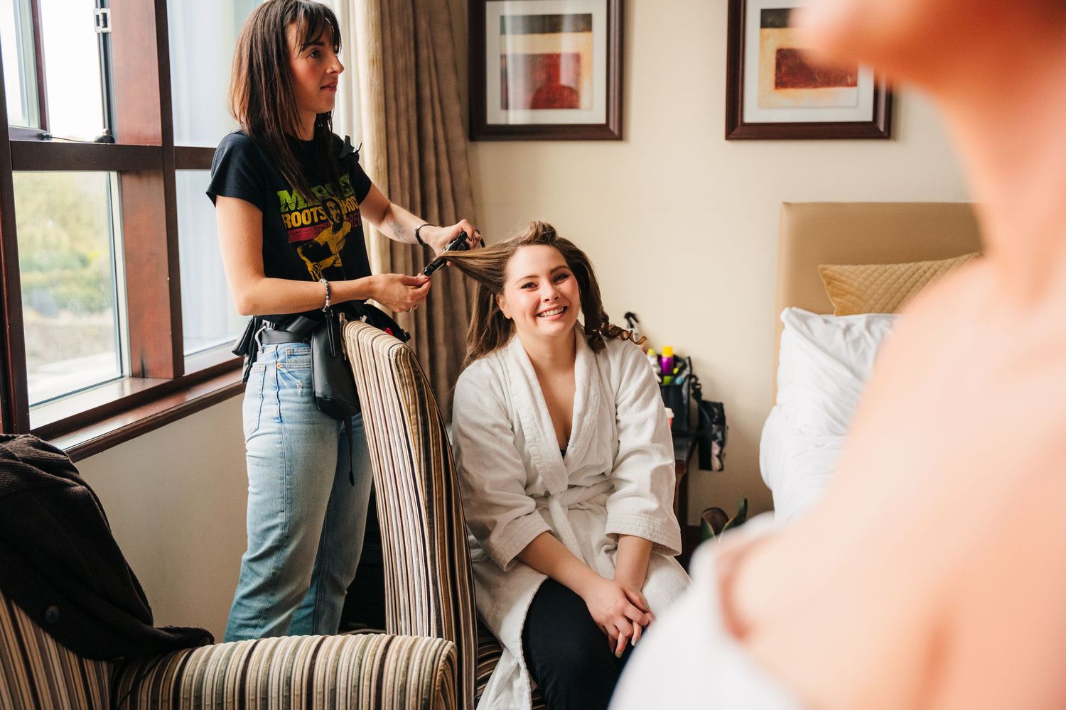 Hair stylist styling hair in a cozy room with natural light from windows and framed artwork on the wall. Wedding Photography Trim Castle Ire