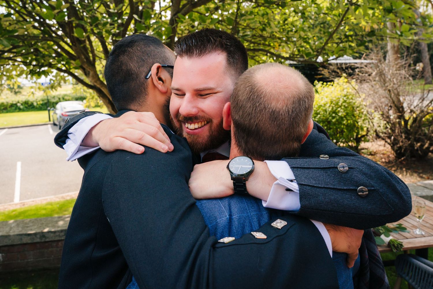 Three people sharing a warm group embrace outdoors on a sunny day. Portmarnock Hotel Wedding PHOTOGRAPHER Dublin