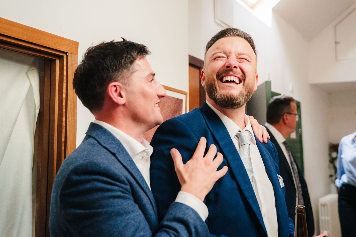 Two people in suits sharing a candid laugh in a bright indoor setting. Same sex wedding PHOTOGRAPHER Dublin