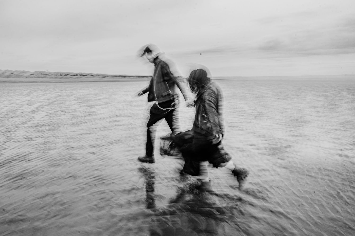 Black and white photo of people wading through shallow water at a beach with reflective surface. engagement PHOTOGRAPHER Dublin