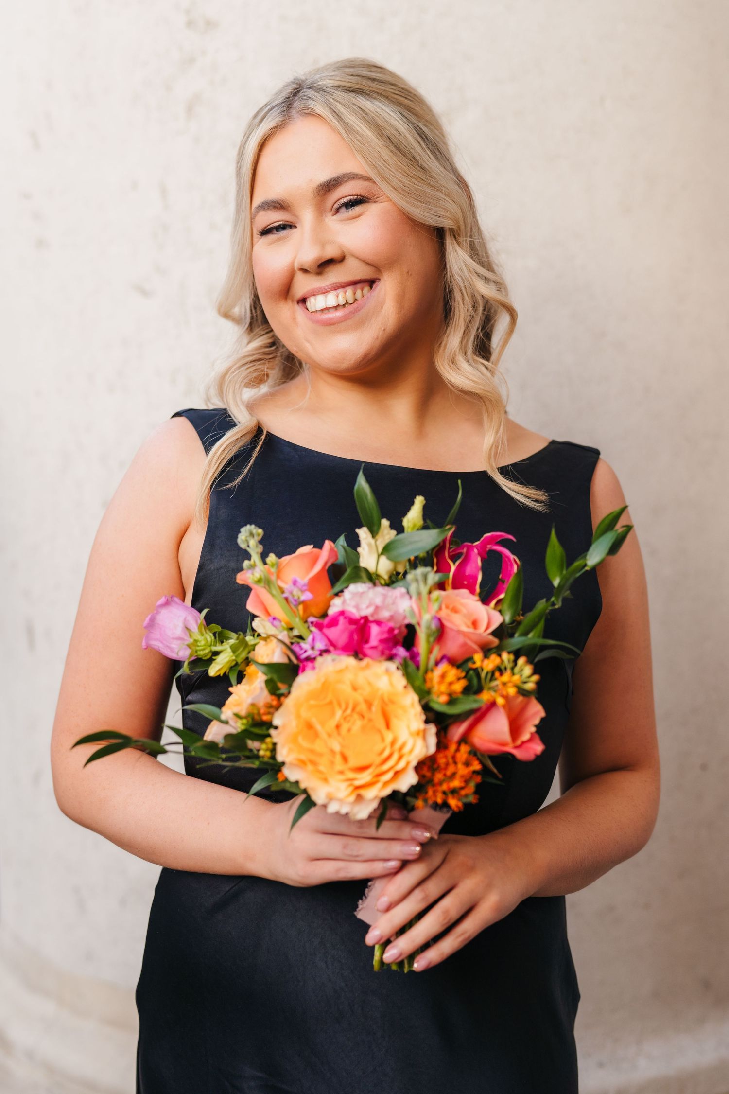 Bridesmaid in black dress holds vibrant orange and pink floral bouquet against neutral backdrop. Wedding photographer dublin