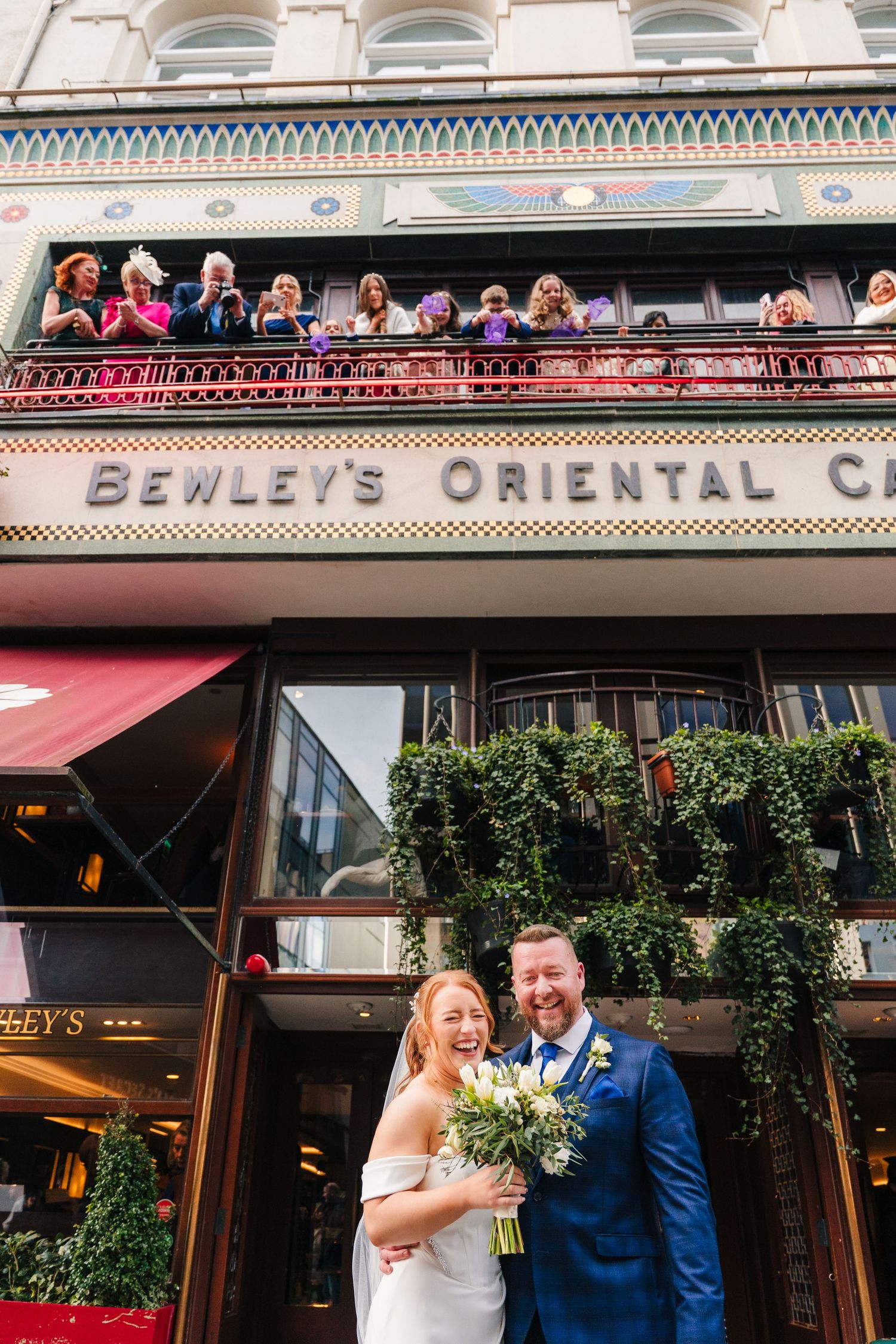 A wedding couple poses in front of Bewley's Oriental Cafe with guests watching from the balcony above. Wedding Photographer Dublin