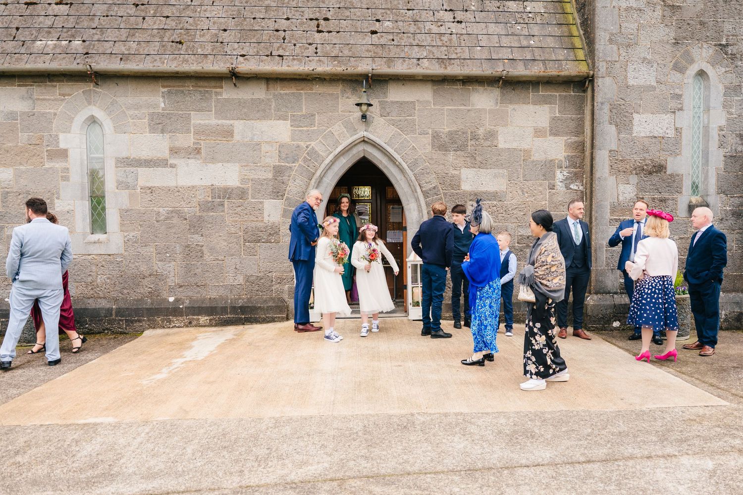 Wedding guests gathered outside stone church building on a sunny day.