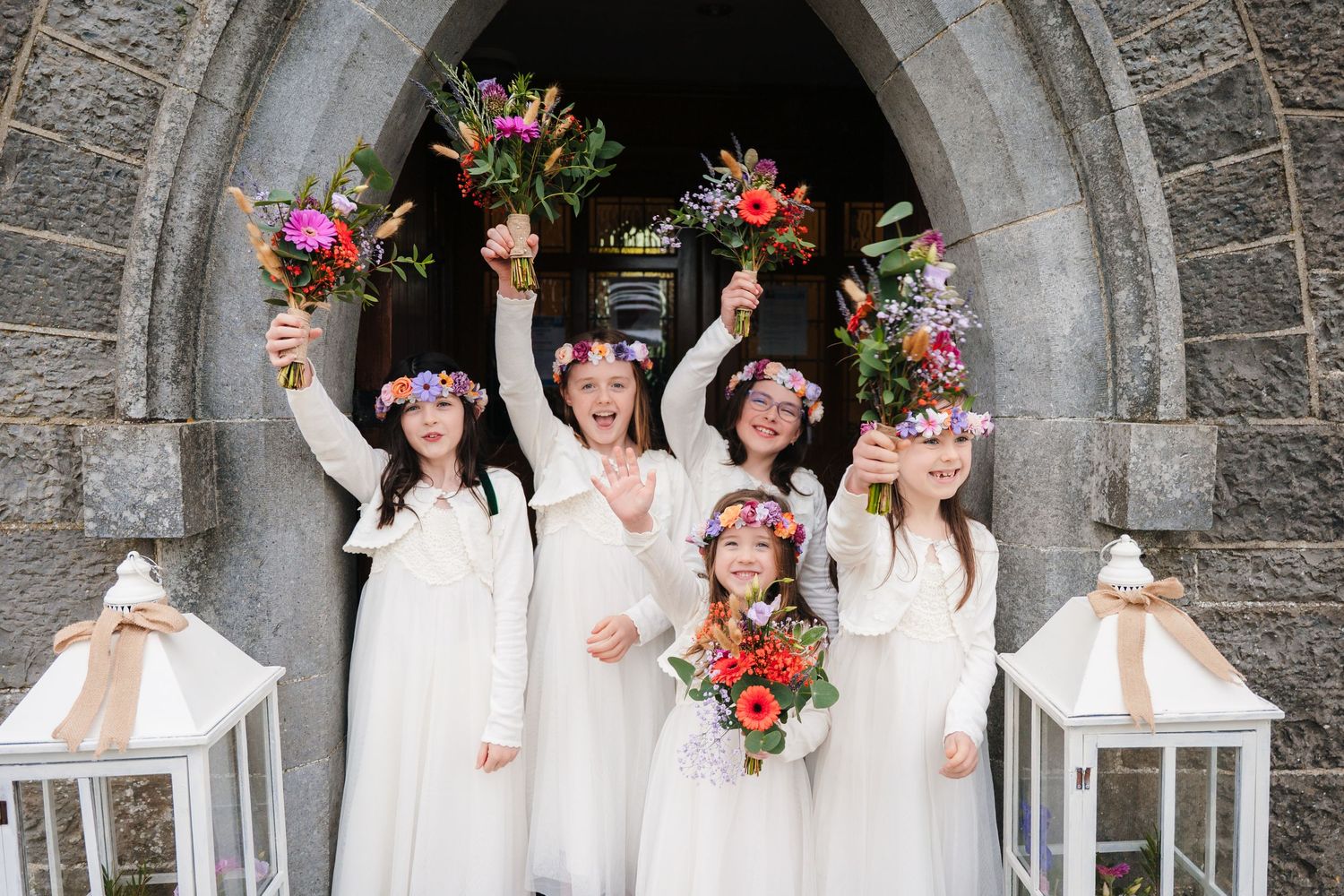 A group in white dresses and floral crowns poses together under a stone archway during a celebration. Wedding PHOTOGRAPHER Dublin Meath