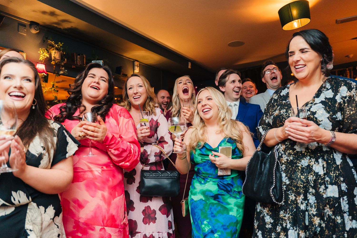 A group of wedding party guests in colorful dresses laughing and enjoying drinks together at a reception. Wedding PHOTOGRAPHER Dublin