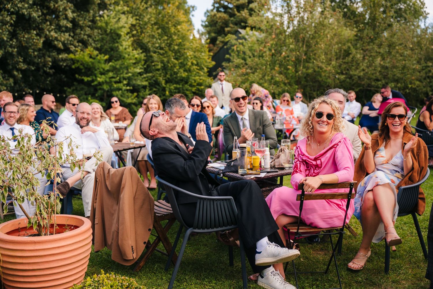 Wedding guests sitting in chairs on a lawn during an outdoor summer ceremony with potted plants and greenery nearby. . Village Barn Tyrrells