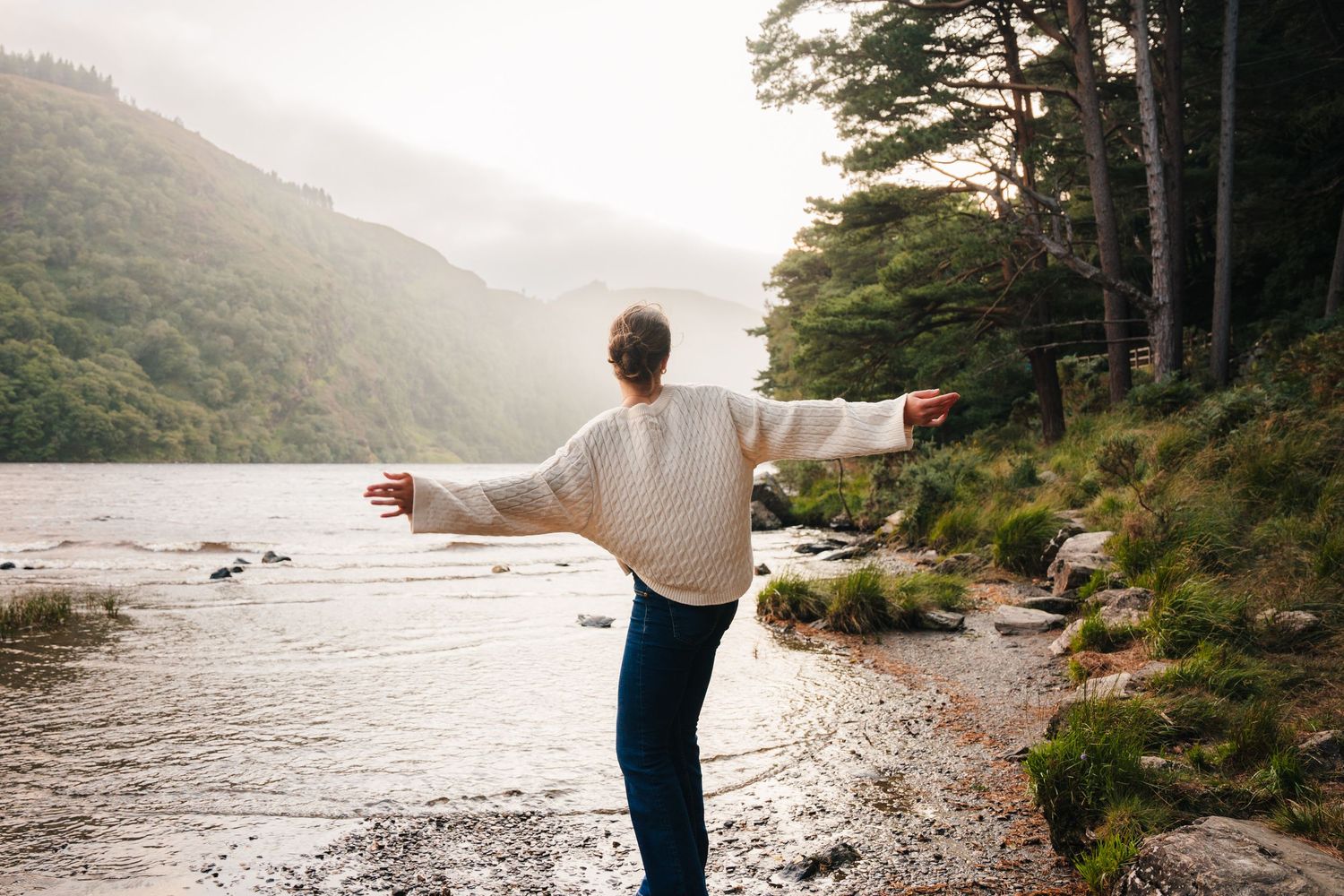 Family photographer Ireland Dublin A person spreads their arms wide while standing at the edge of a mountain lake surrounded by forest.