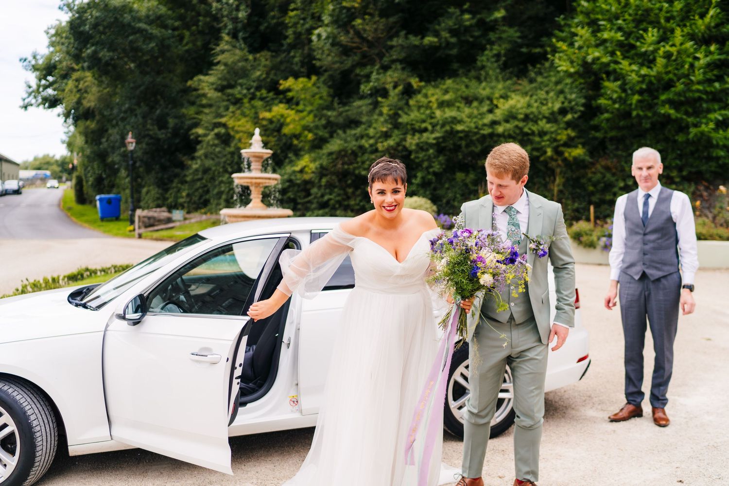 Millhouse slane wedding photographer dublin Wedding couple exits white car as groomsman in grey suit follows behind on outdoor path.