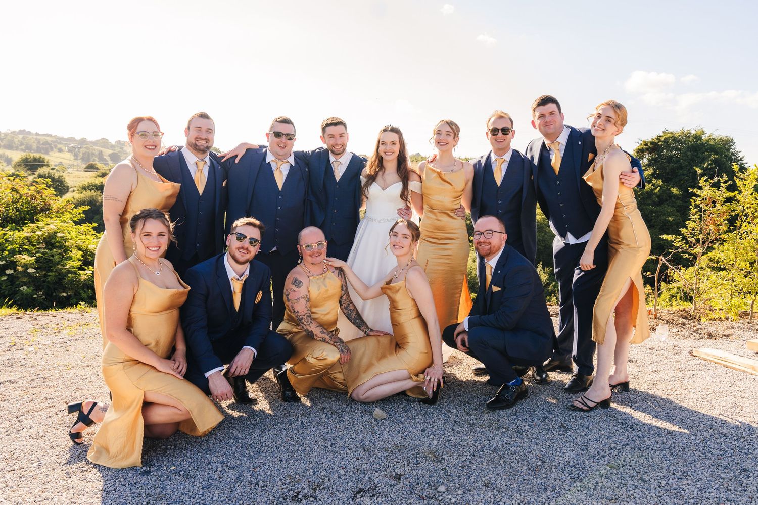 Poulaphouca Wedding photographer wicklow  Wedding party poses together on hillside wearing navy suits and gold bridesmaid dresses.