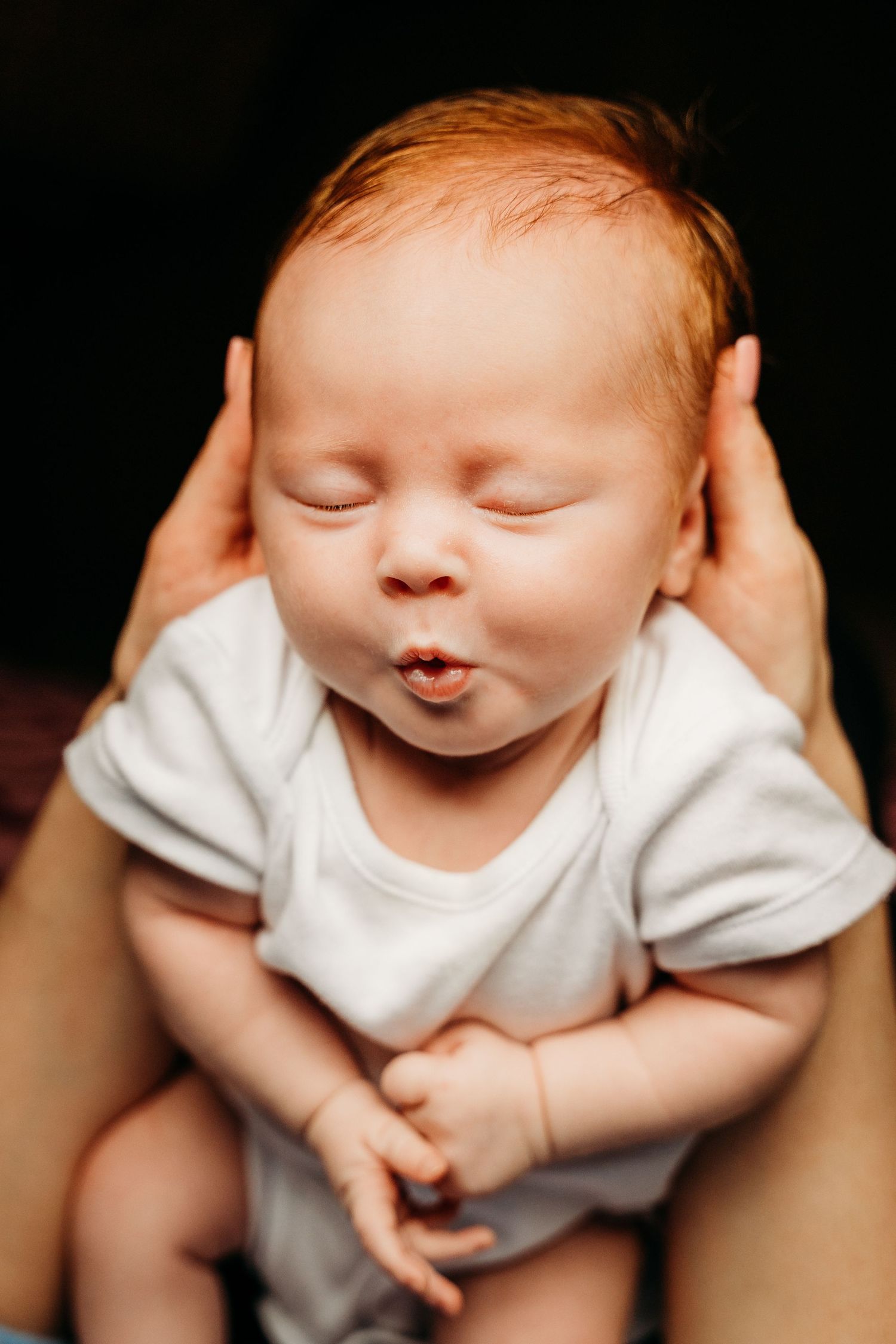Peaceful sleeping newborn baby wearing white onesie being cradled by gentle hands against dark background. Family PHOTOGRAPHER Dublin