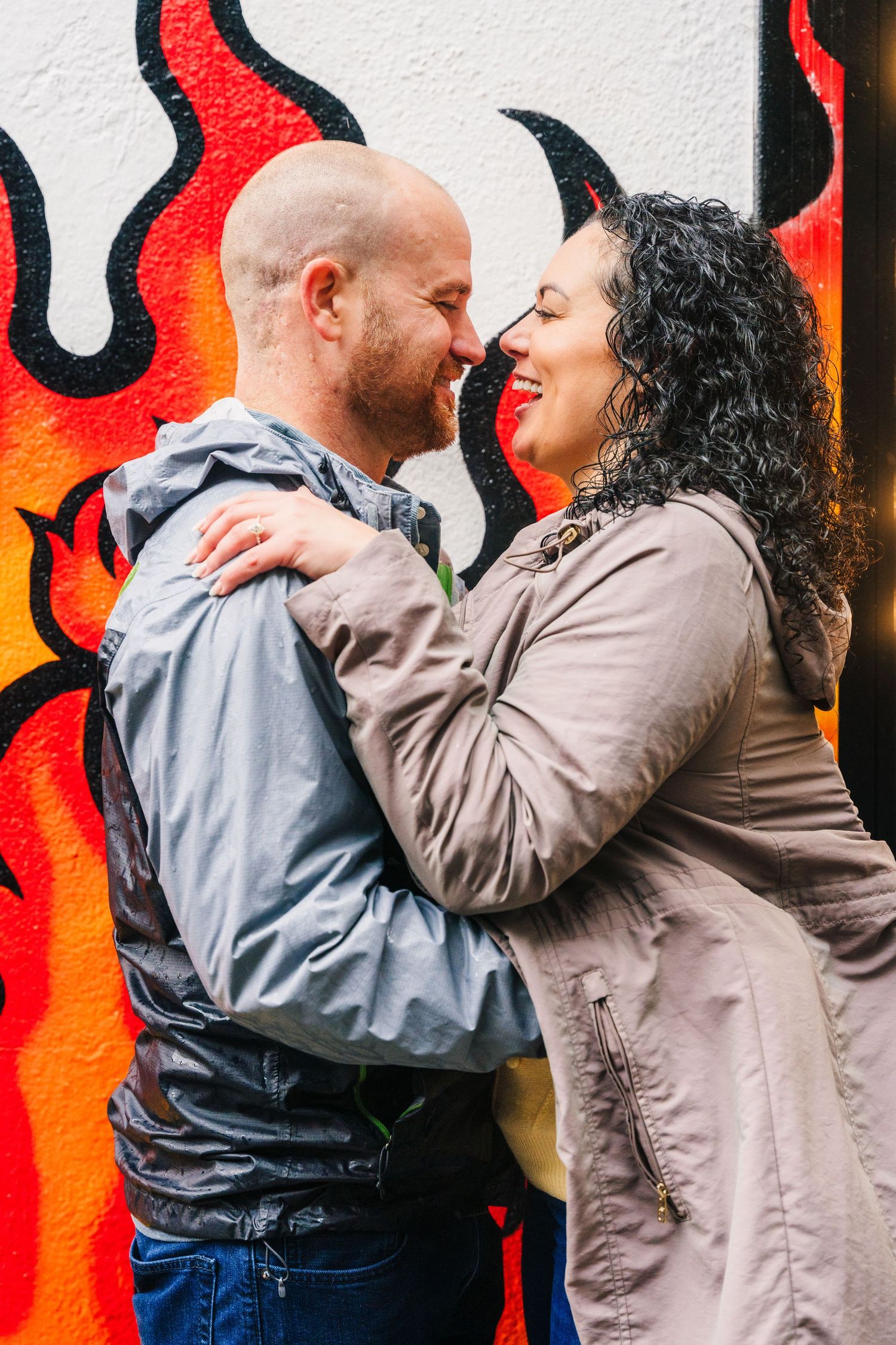 A loving couple embraces while standing in front of a vibrant red and orange flame mural painted on a white wall. Proposal Dublin Photograph