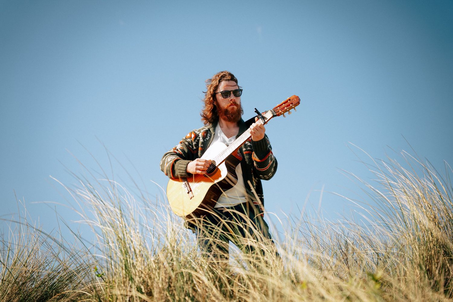 A musician plays acoustic guitar while sitting in tall beach grass with blue sky in the background. PHOTOGRAPHER Dublin