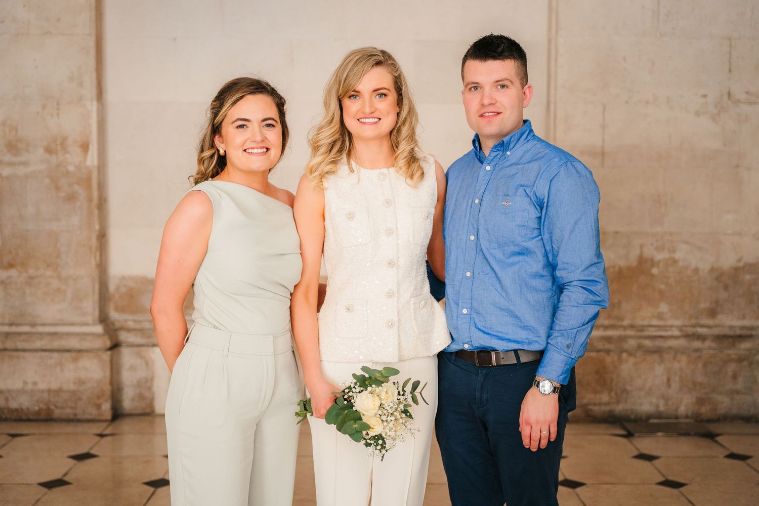 Three people in formal attire pose together against neutral wall with white bouquet. City Hall Dublin wedding photogrAPher