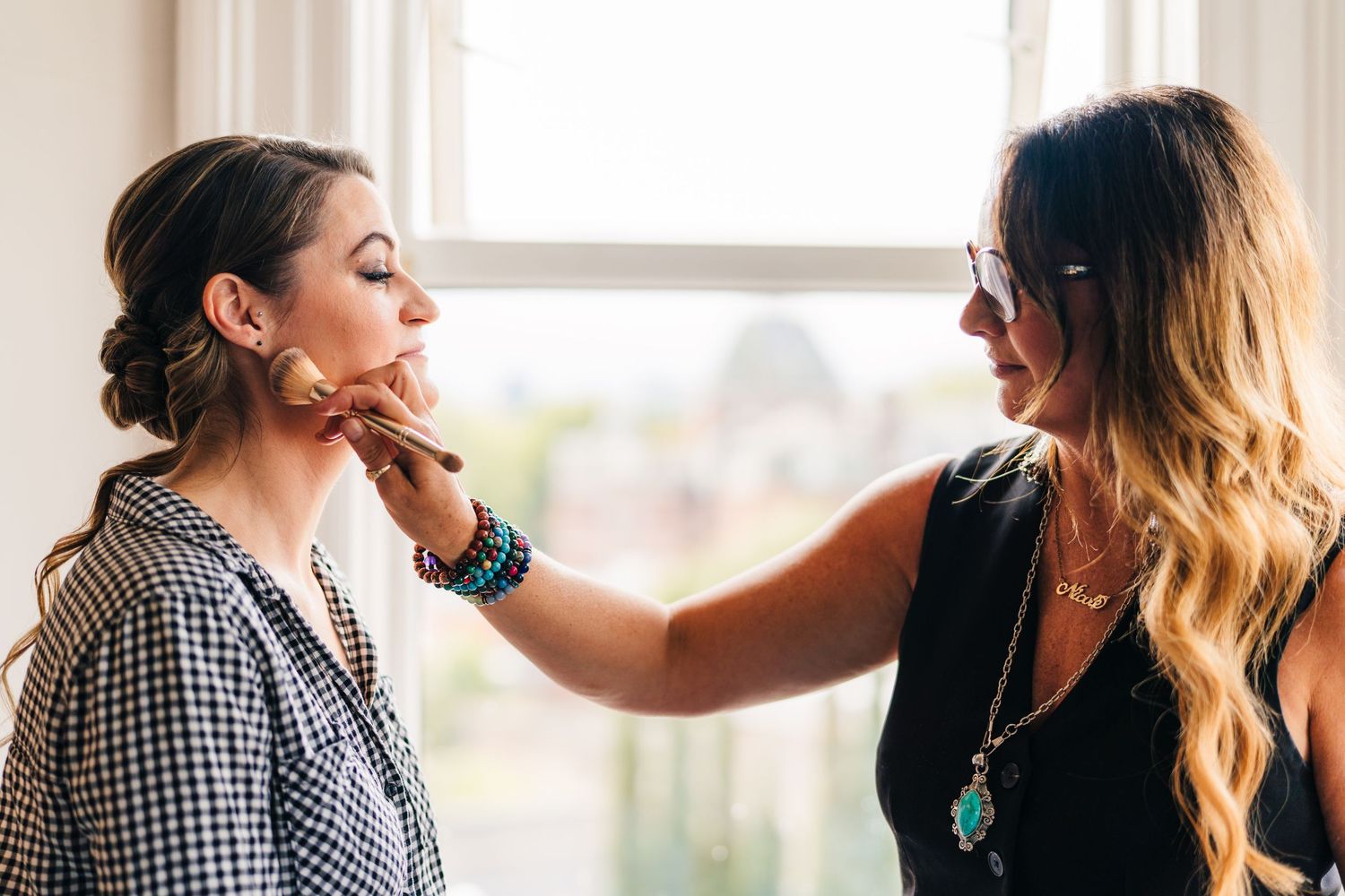 Professional stylist applying makeup artistry while preparing model for photoshoot in sunlit studio. House Dublin Wedding Photographer