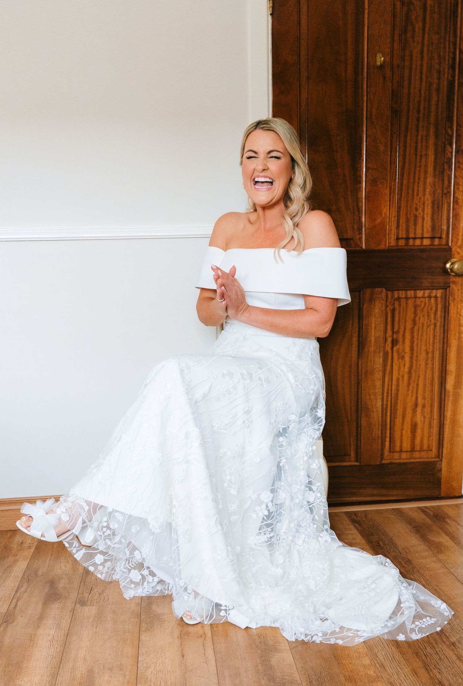A bride in a white lace wedding dress sitting and laughing joyfully on a wooden floor. The Dean Dublin Wedding Photographer