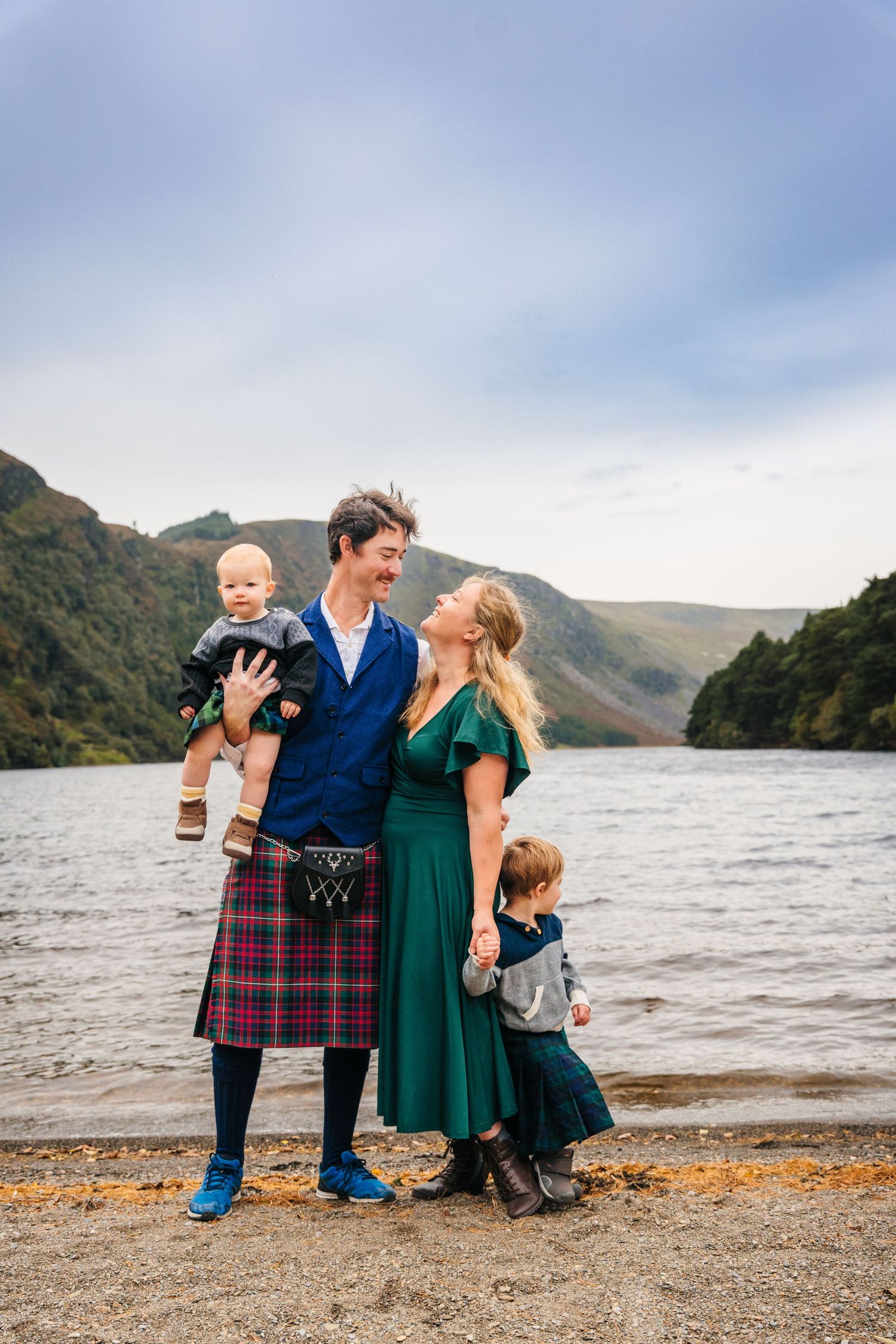Family in coordinated outfits stands together on a rocky lakeshore with mountains in the distance. Family photogrAPHER DUBLIN