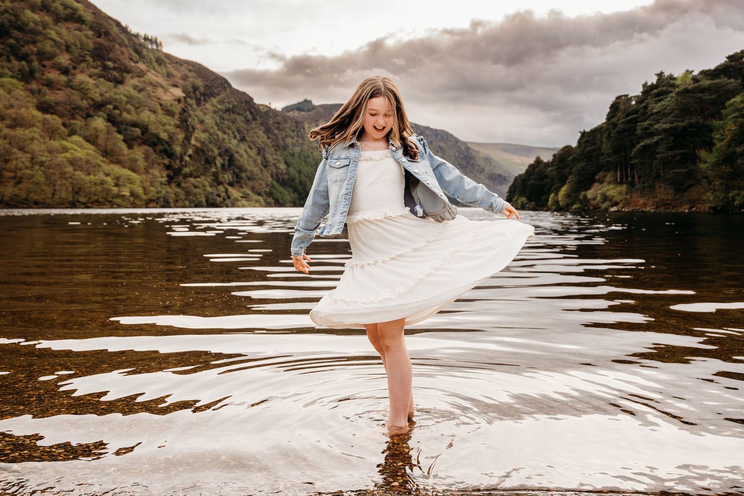A dancer in a white dress and denim jacket spins in mountain lake waters during a moody sunset. family PHOTOGRAPHER Dublin