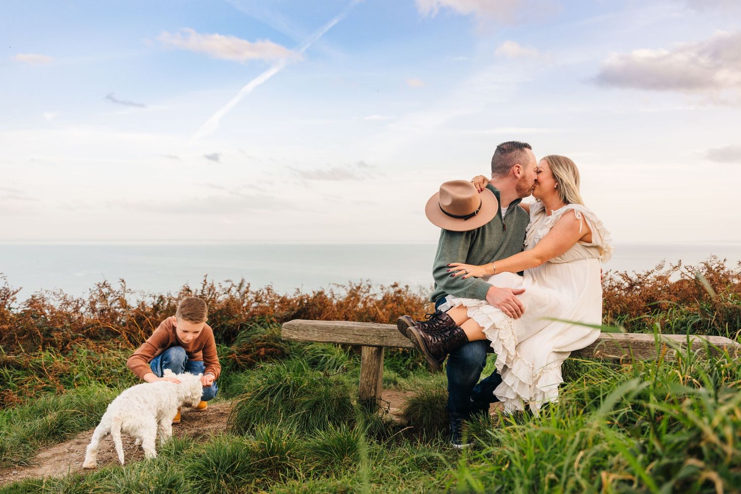 Couple shares a romantic moment on wooden bench at scenic coastal overlook while photographer captures the scene with dog nearby. family PHO