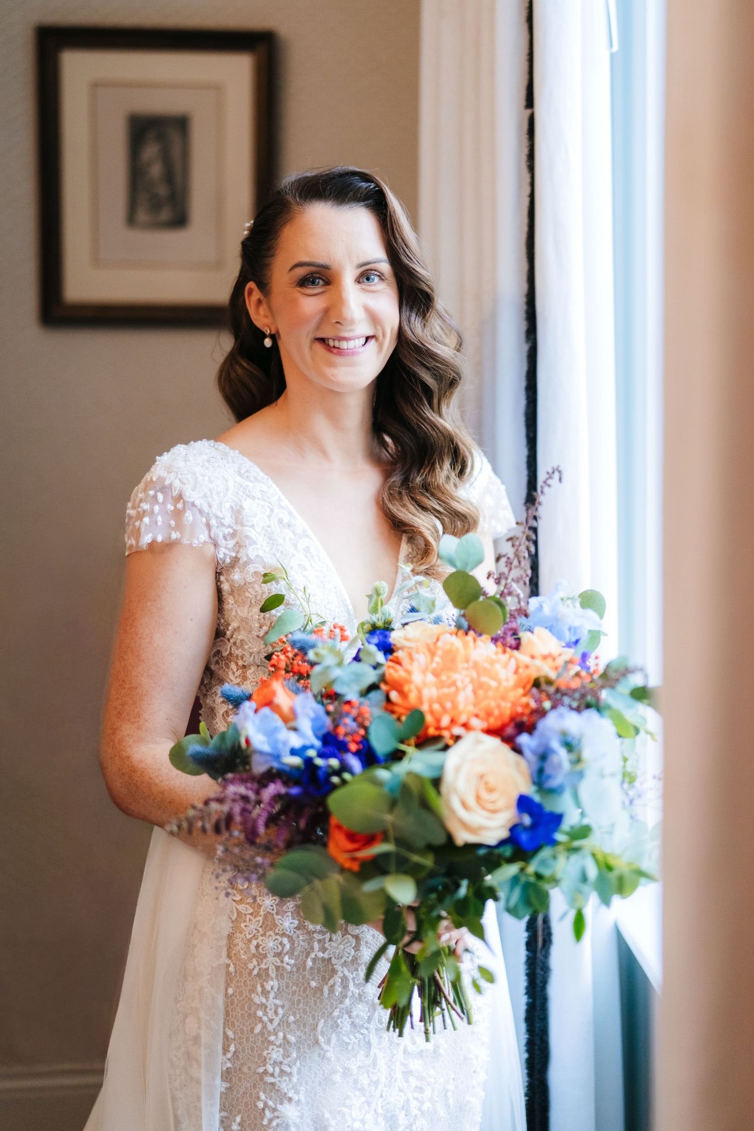 A bride in a beaded wedding gown holds a vibrant bouquet of blue, peach and orange flowers by a window.