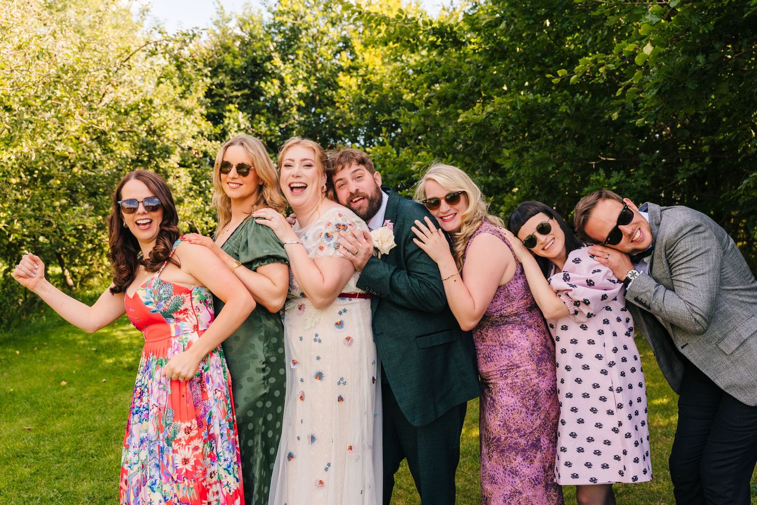 Wedding party poses together playfully in a garden setting wearing colorful dresses and formal attire. . Village Barn Tyrrellspass wedding p