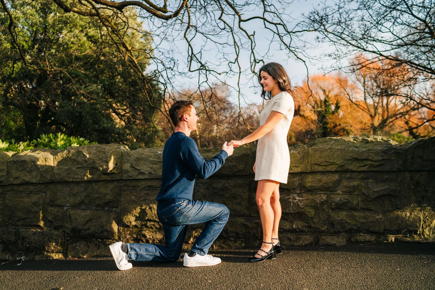 A romantic proposal moment takes place outdoors against a rustic stone wall during golden autumn weather. Surprise proposal dublin photograp