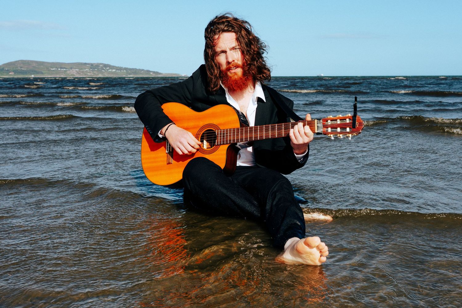 Musician sits in shallow ocean water playing acoustic guitar with mountains visible in background. PHOTOGRAPHER Dublin
