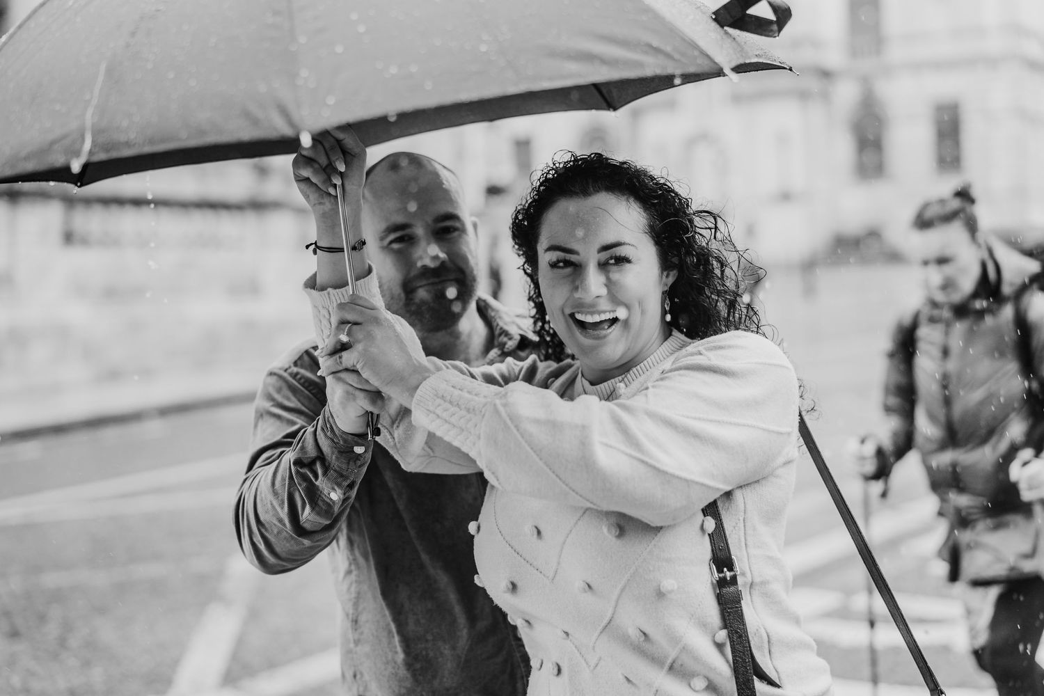Black and white image of joyful couple laughing together under umbrella by stone architecture. Proposal Dublin Photographer