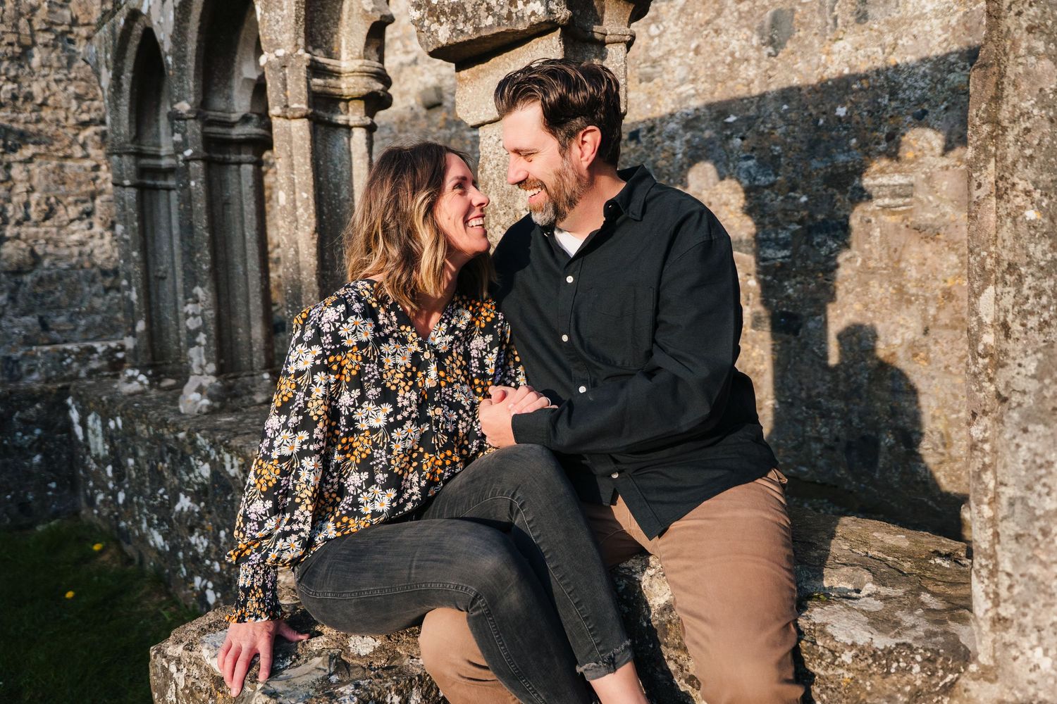 A couple shares a sweet intimate moment while sitting against an old stone wall in warm evening sunlight. Photographer Ireland dublin