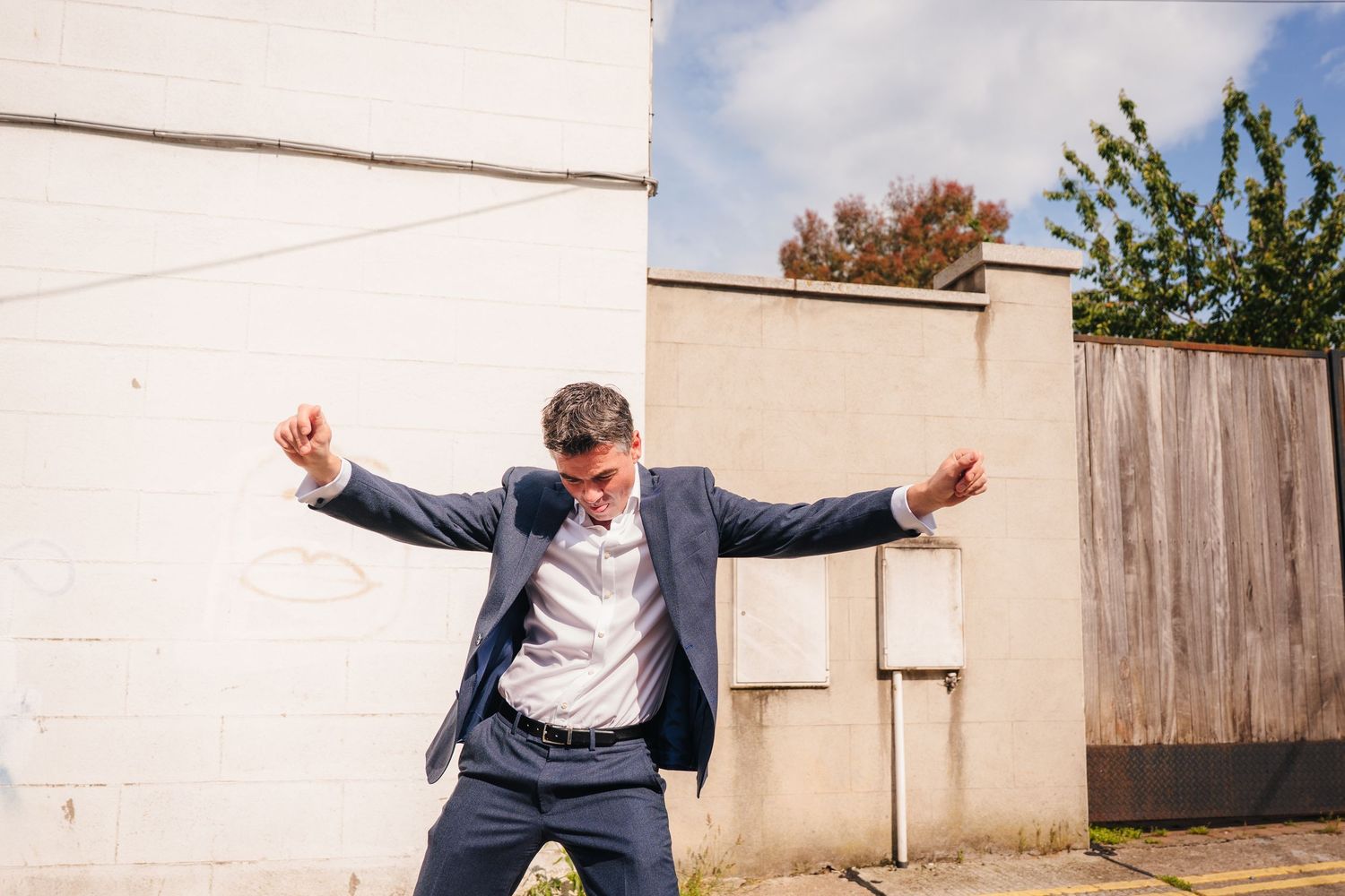 A person in a navy suit striking an energetic pose against a white wall. Same sex wedding PHOTOGRAPHER Dublin