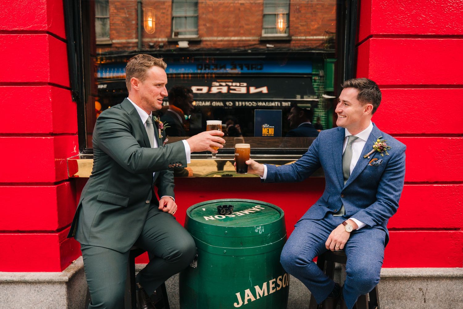 Two people in formal attire sharing drinks and conversation in front of a bright red wall and green bar furniture. Same sex wedding PHOTOGRA