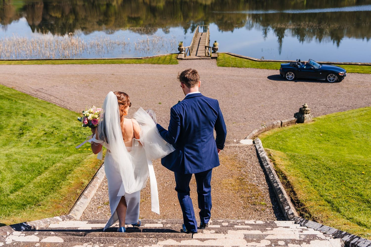 Couple walks down steps near serene lake with green grass and reflection of landscape on water. Castle Leslie Wedding Photographer Dublin