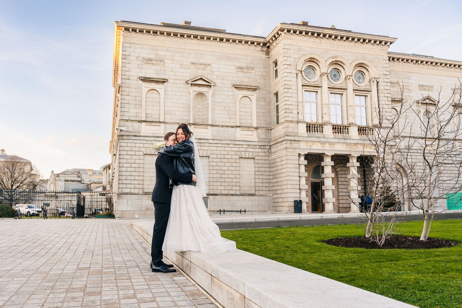 A wedding couple embraces outside a grand historic stone building with arched windows. Wedding PHOTOGRAPHER Dublin