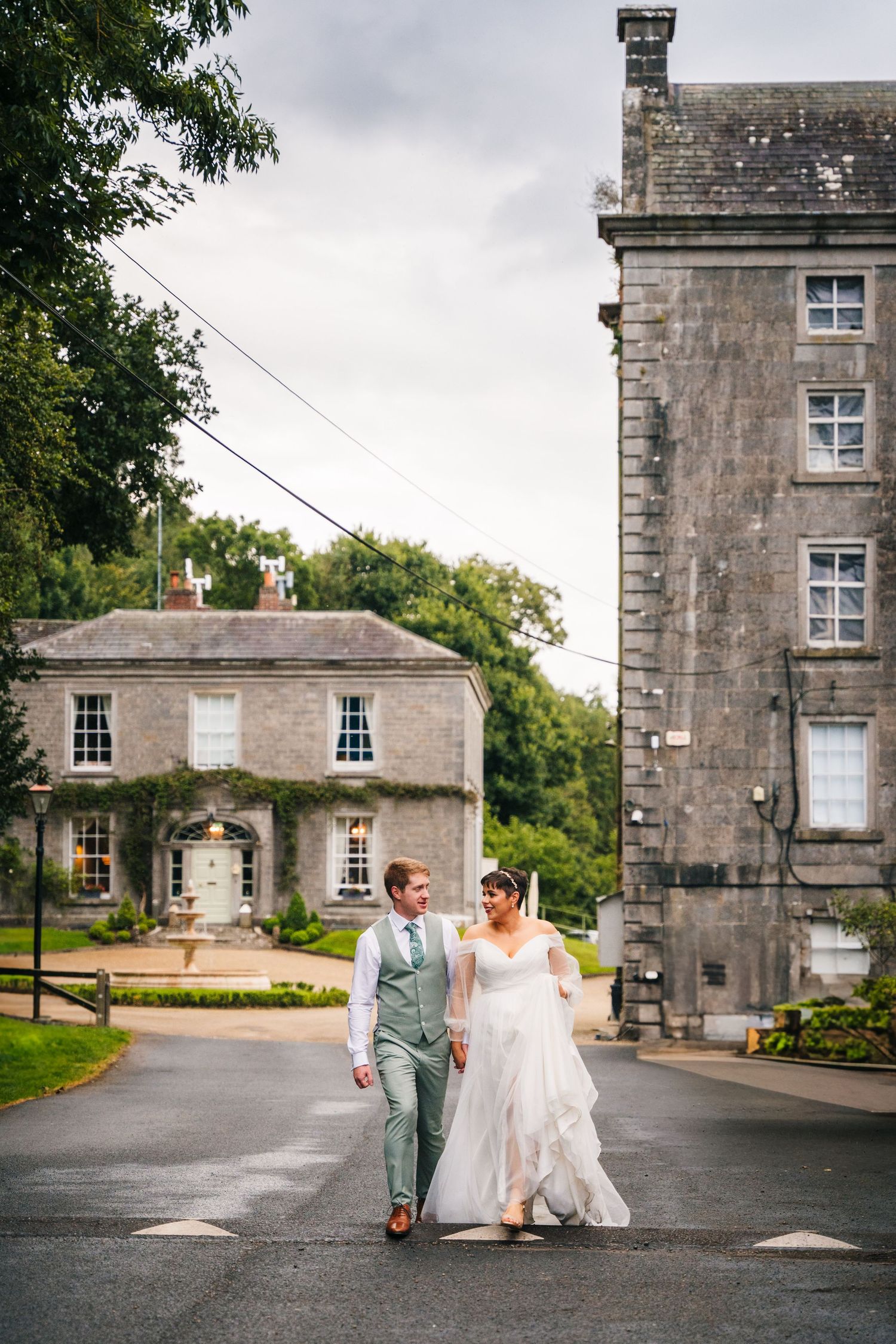 Millhouse slane wedding photographer dublin Newlyweds walk together on path between historic stone buildings on overcast day.