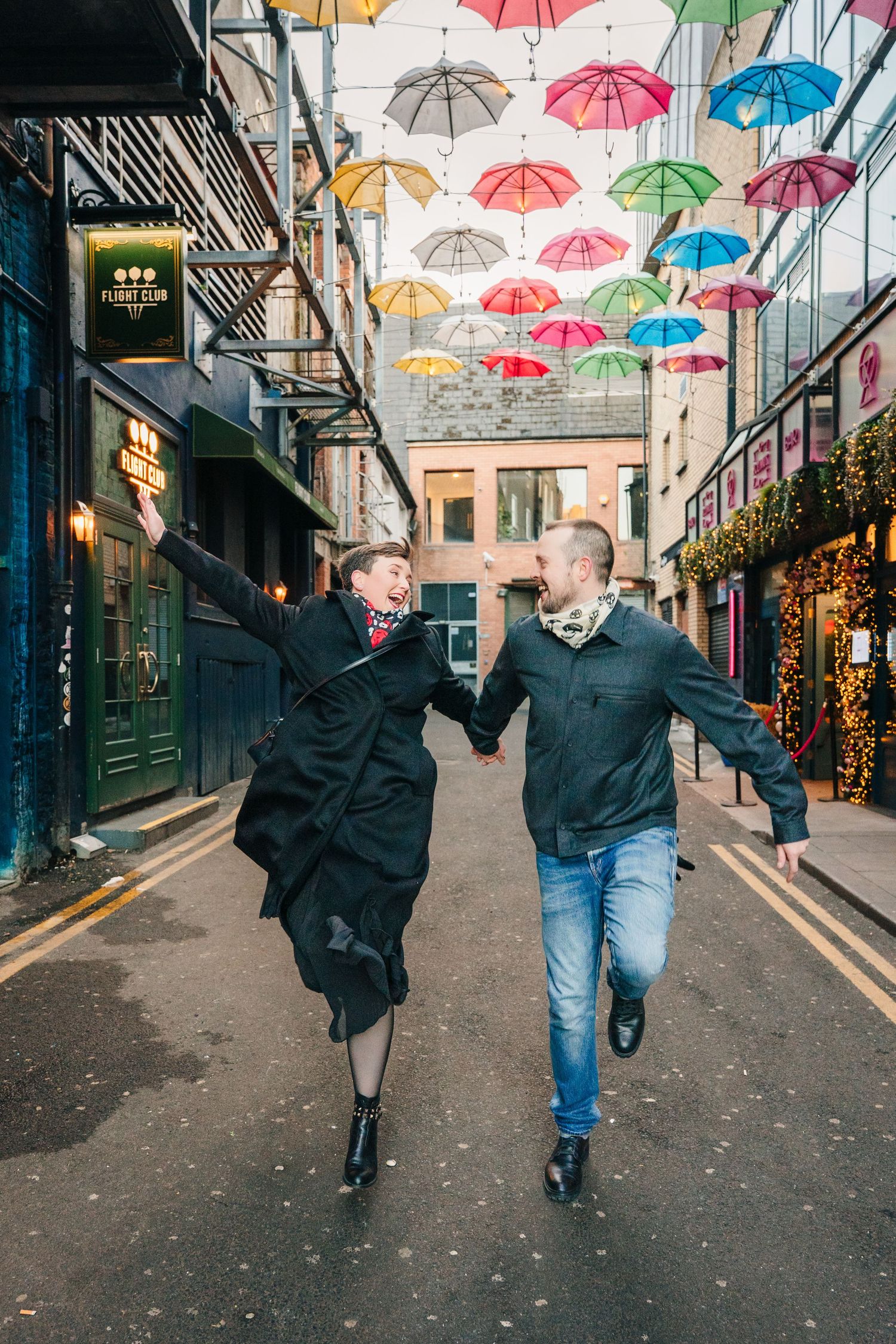 Two people walk under a colorful umbrella art installation in an urban alleyway decorated with string lights. family PHOTOGRAPHER Dublin