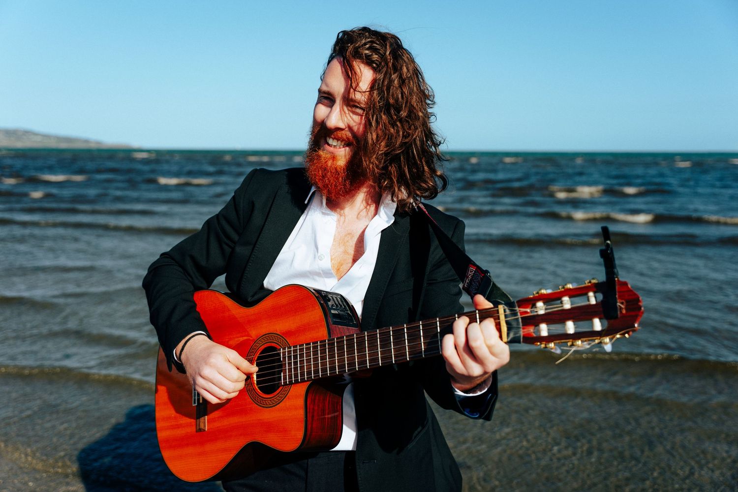 Musician with long red hair and beard plays acoustic guitar while sitting on beach. Professional headshots PHOTOGRAPHER Dublin