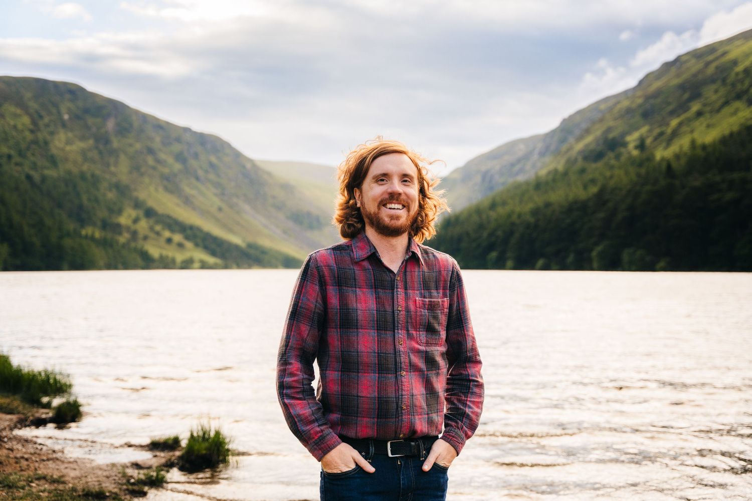 Person in plaid flannel shirt stands at mountain lake with scenic valley and peaks in background. Professional headshots PHOTOGRAPHER Dublin