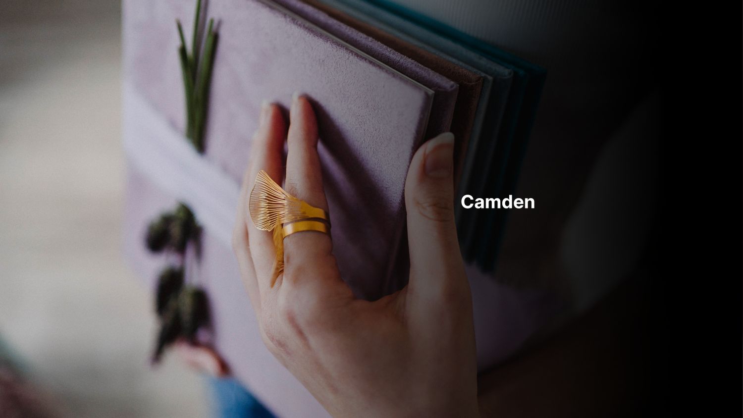 Close-up of a hand wearing a gold Camden ring, gently holding an open floral book.