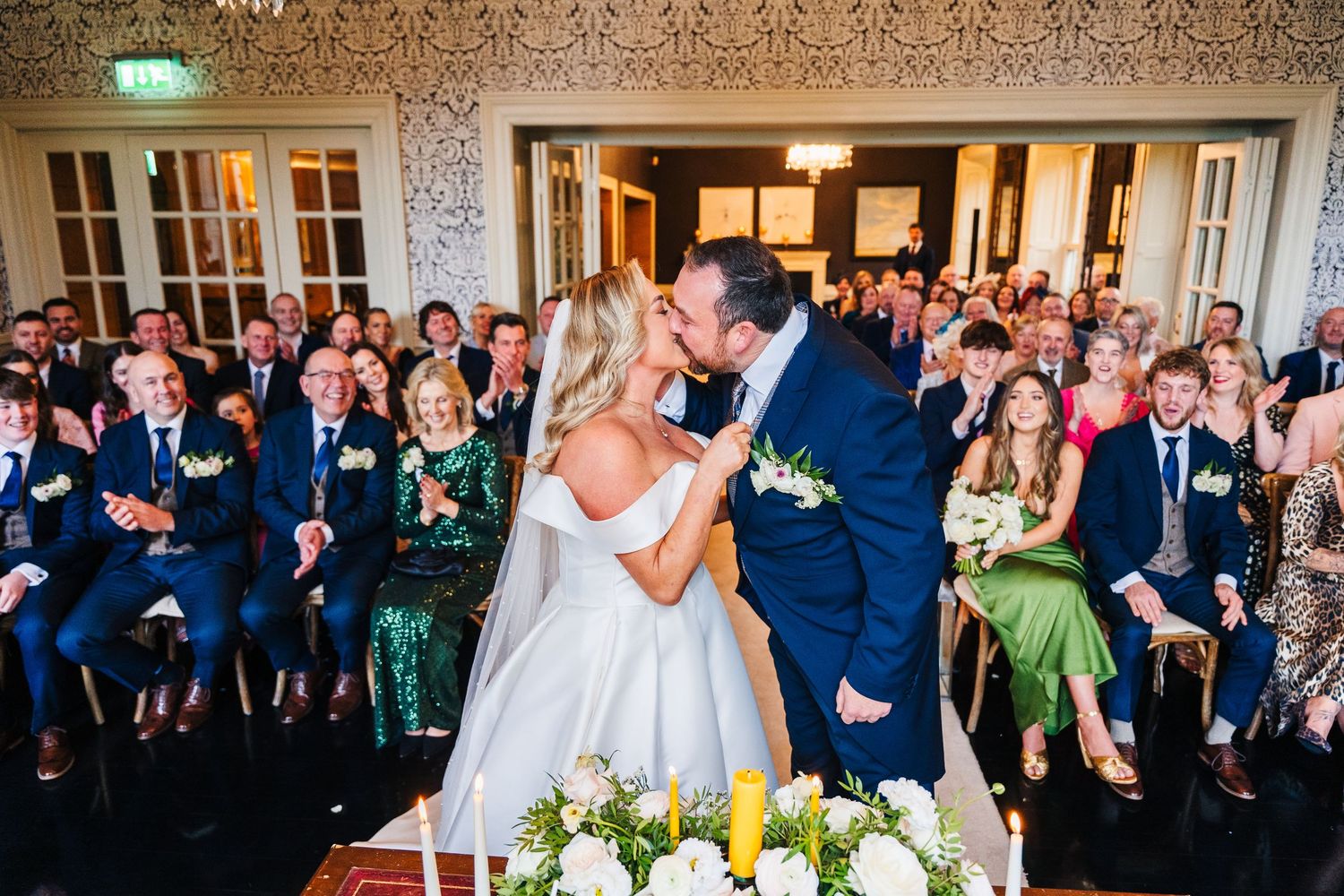 Couple shares a romantic kiss at their wedding ceremony while guests look on inside an elegant venue with ornate walls.  Virginia Park Lodge