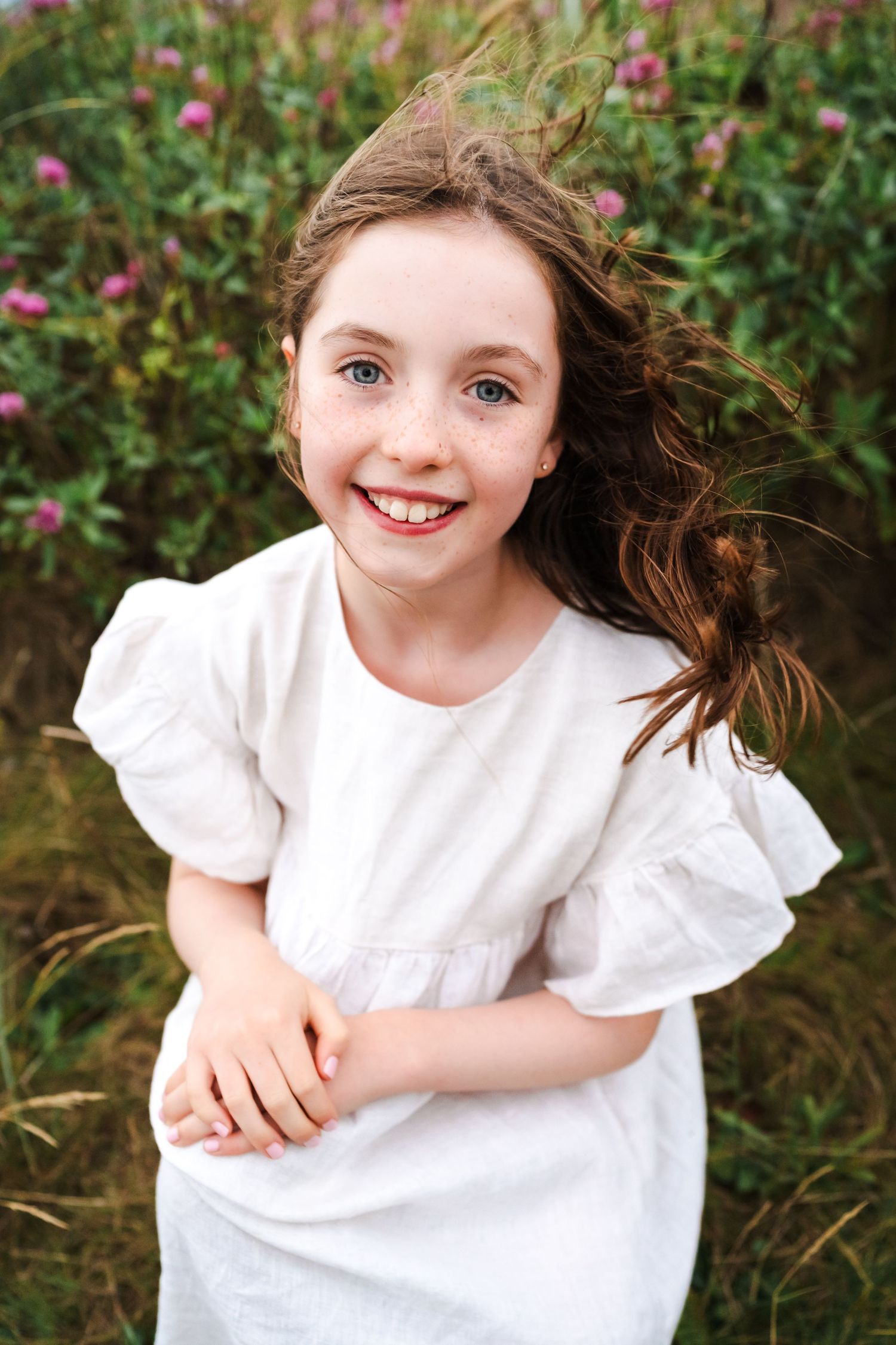 Young girl in white dress smiles brightly among pink wildflowers on a sunny day.  Family photographer Dublin Ireland