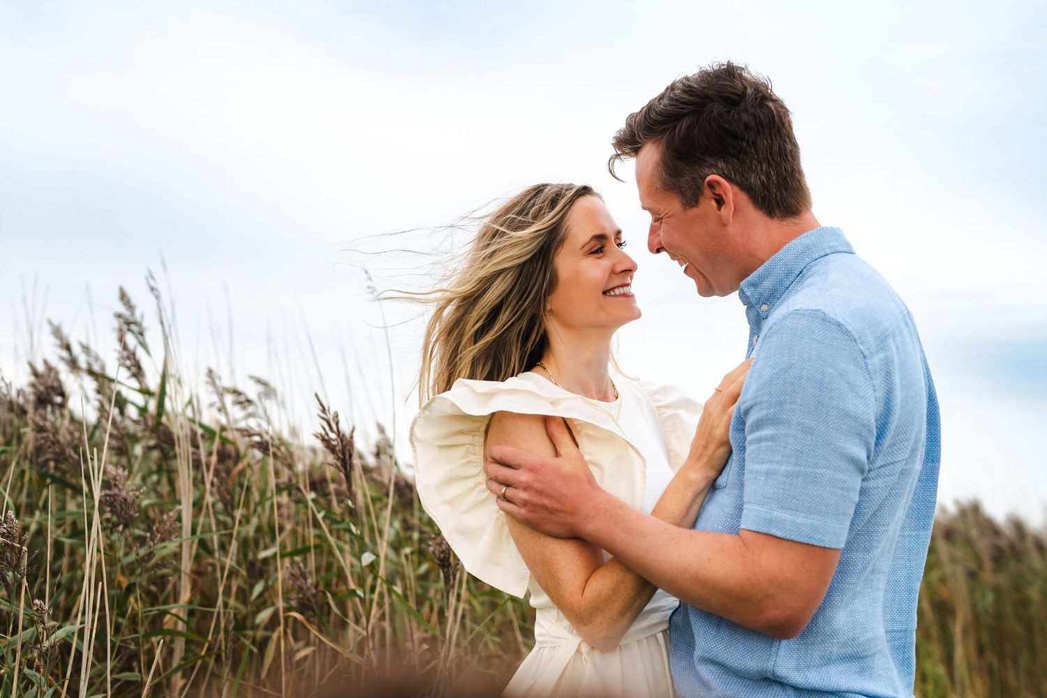 A couple embraces in a field of tall grass with their hair blowing in the wind against a cloudy sky.  Family photographer Dublin Ireland