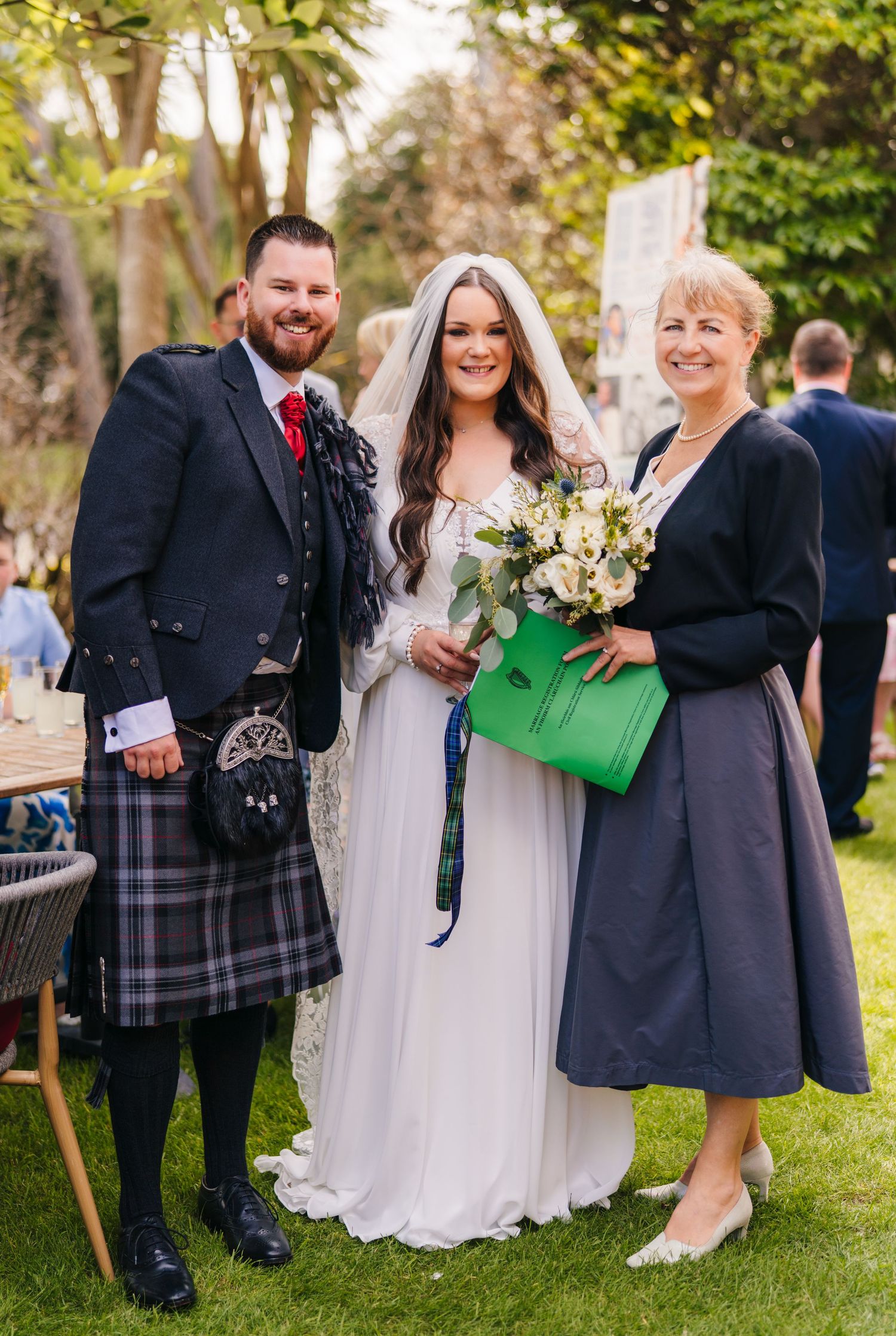 A wedding party in Scottish attire poses outdoors with the bride in white and guests wearing traditional Highland dress. Portmarnock Hotel W