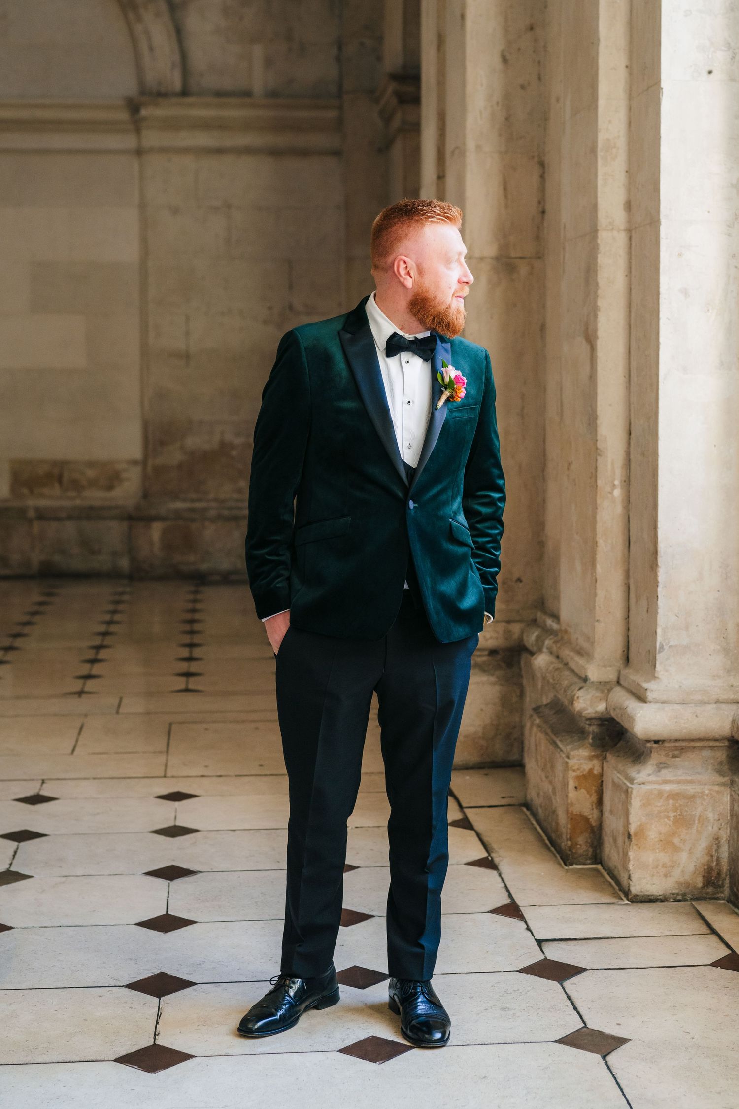 Groom in emerald velvet tuxedo and bow tie poses against ornate marble columns with checkered floor. City Hall Dublin Photographer