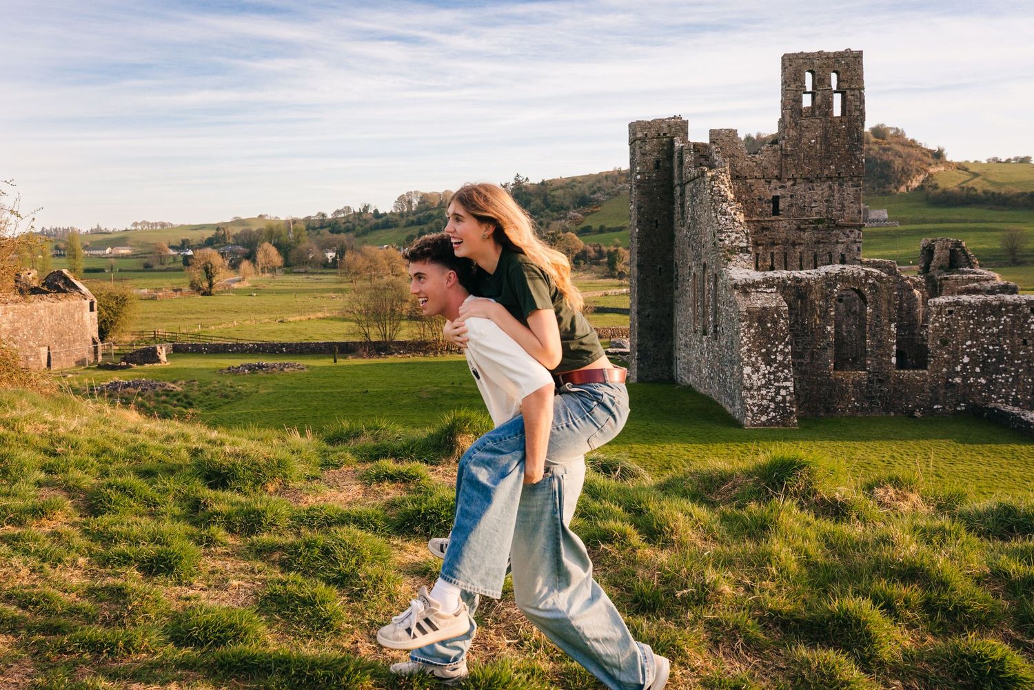 Two people playfully posing on grassy hillside with ancient castle ruins in background. Photographer Ireland dublin