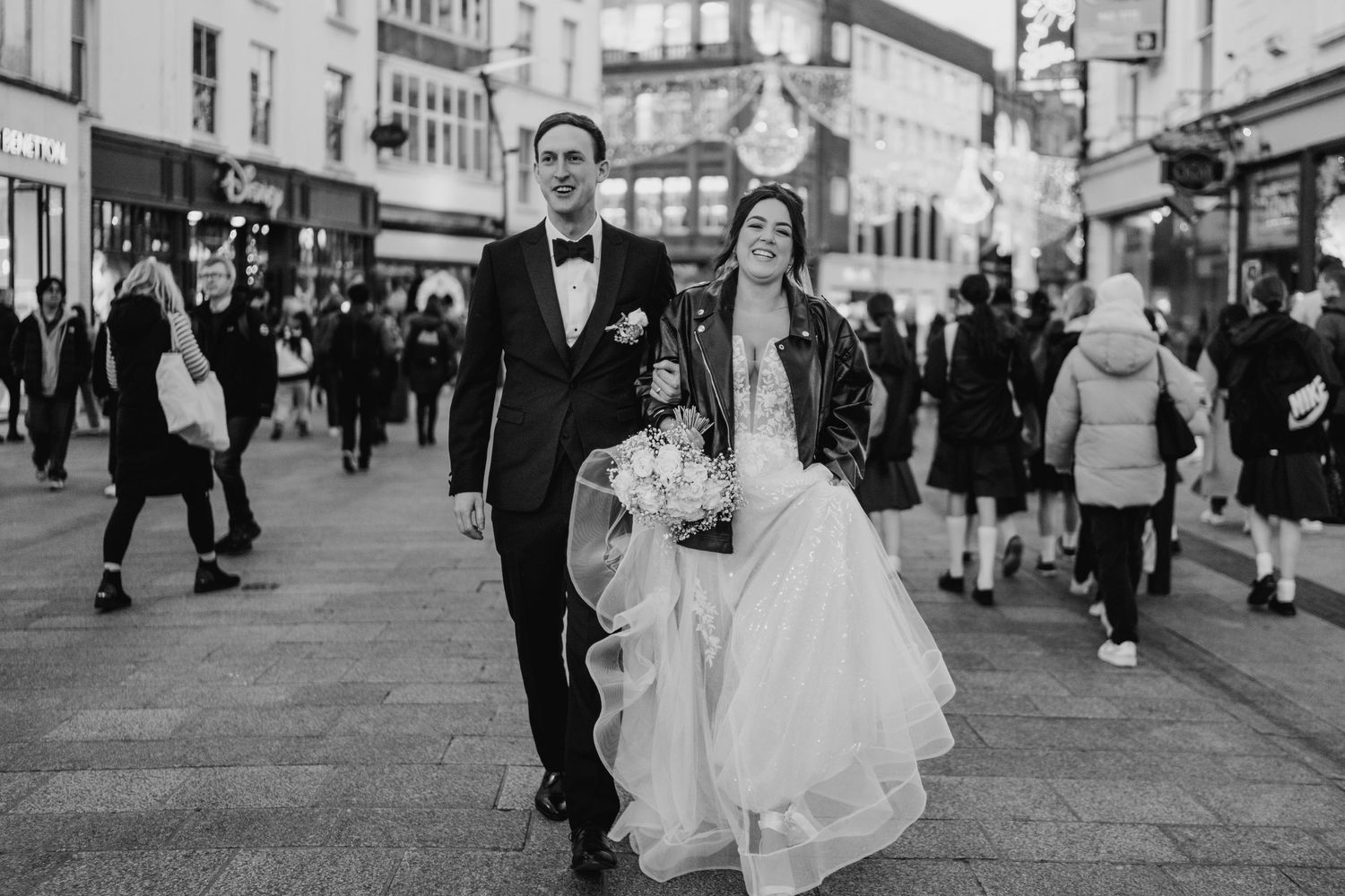 A black and white photo of a couple walking down a busy pedestrian street in formal attire. Wedding PHOTOGRAPHER Dublin