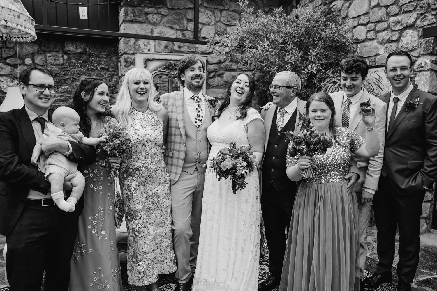 Wedding group photo with bridal party in floral dresses against stone wall backdrop. Wedding photographer Dublin