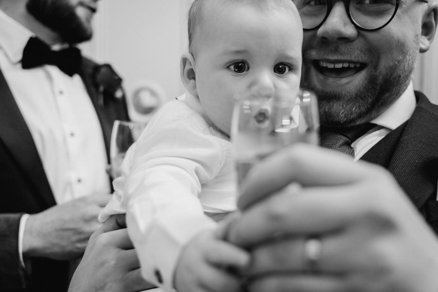 Close-up black and white photo of a baby in formal wear reaching out at a celebration.