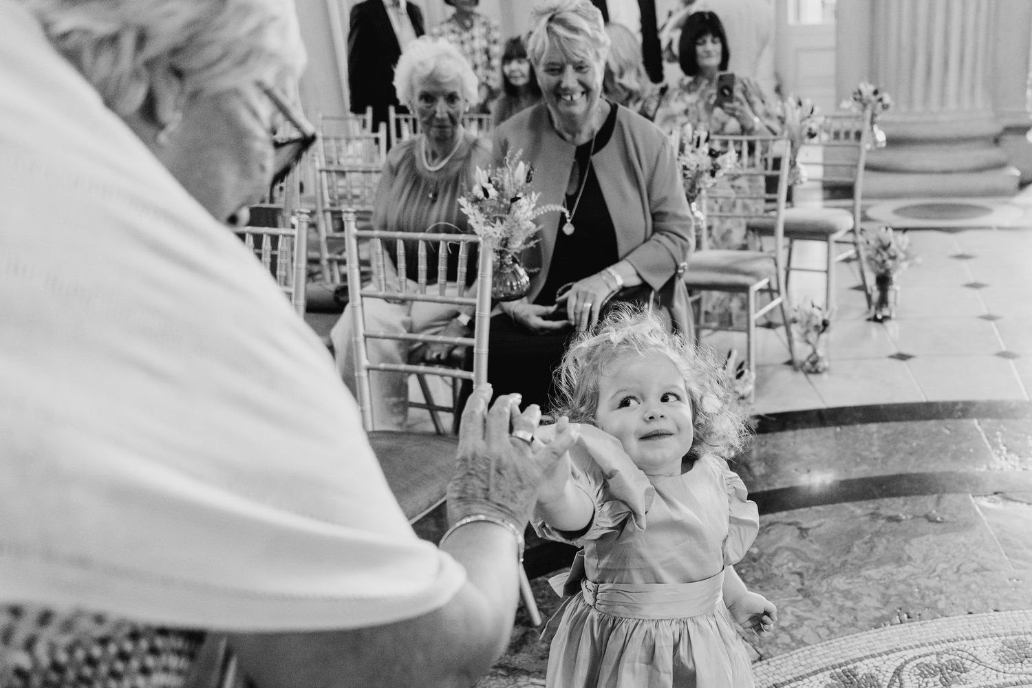 A small child in a white dress dances joyfully at an elegant event while adults watch from nearby chairs.  city hall dublin wedding photos